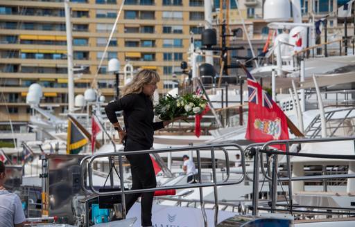 Female dressed in black standing on the bow of a superyacht at the Monaco Yacht Show, Monte Carlo visible in background.