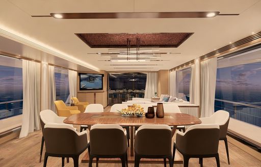 wooden dining table surrounded by brown and white chairs in the window-lined main salon of FD92 yacht