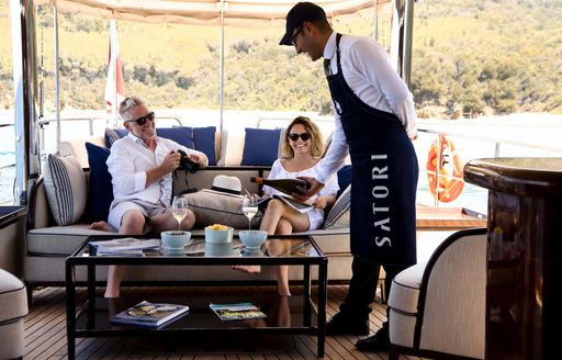 Male crew member pouring drink for two guests on sofa