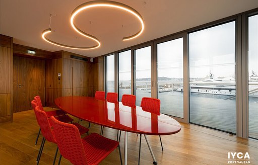 Overview of a conference room at the International Yacht Club d’Antibes (IYCA), long red oval table surrounded by chairs, with large windows in the background.