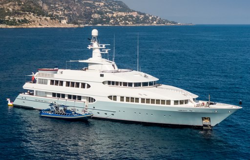 Feadship yacht OLYMPUS on anchor in calm sea on bright day, yacht tender berthed alongside starboard side, land in background