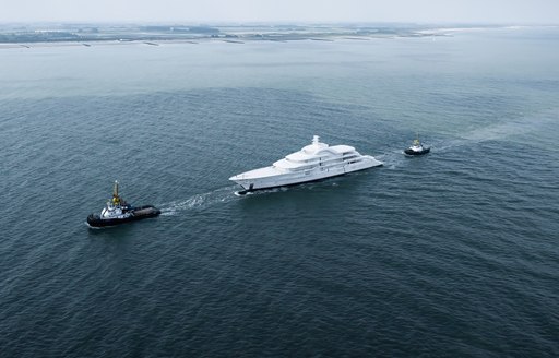 Amels 120 being escorted by tug boats on route to Vlissingen, surrounded by water with land in far distance.