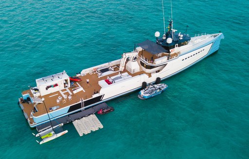 Damen Yacht Support 5009 SHADOW aerial view over starboard quarter deck and side profile, stationary in calm waters, all shell doors appear to be open, with tenders and watercraft moored alongside