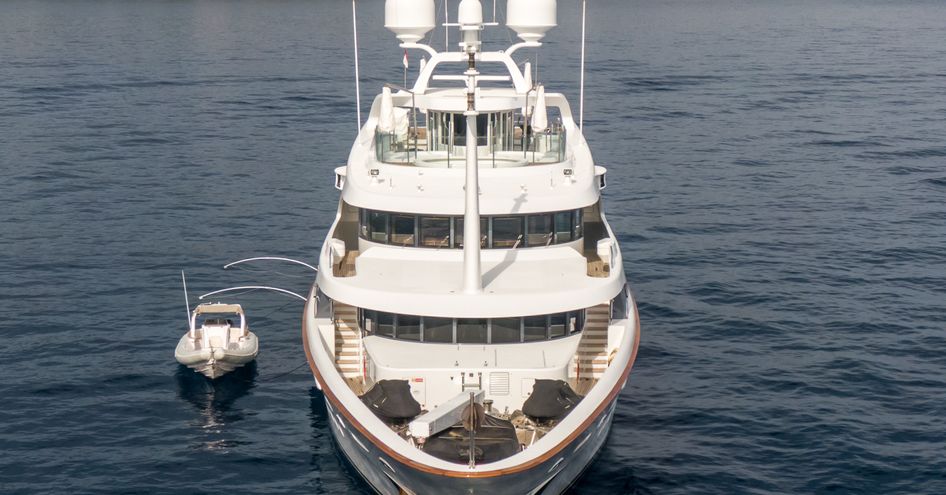 Forward shot of M/Y MEAMINA bow with yacht at anchor. Surrounded by sea with cliff face in background and a small tender to port side.