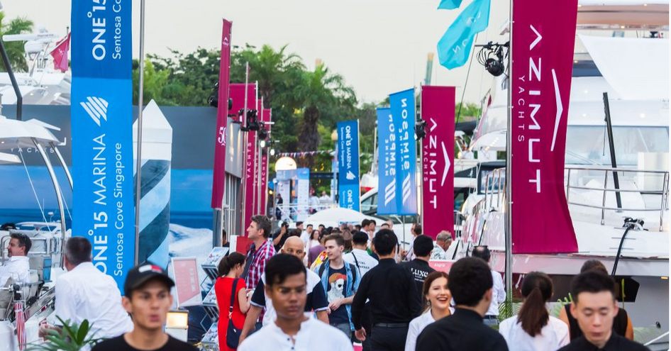 Flags and exhibitors at Singapore Yacht Show, visitors walking along pontoon in between yachts on both sides.