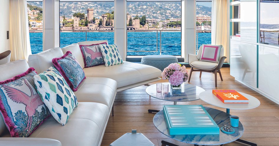 Main salon onboard CRN RIO superyacht, white sofas and vibrant cushions face flatscreen TV, full length windows in background.