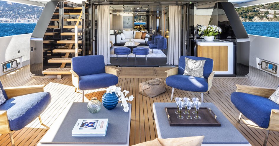 Aft deck onboard Custom Line 140. Low coffee tables surrounded by blue seating, interior visible through open doors.