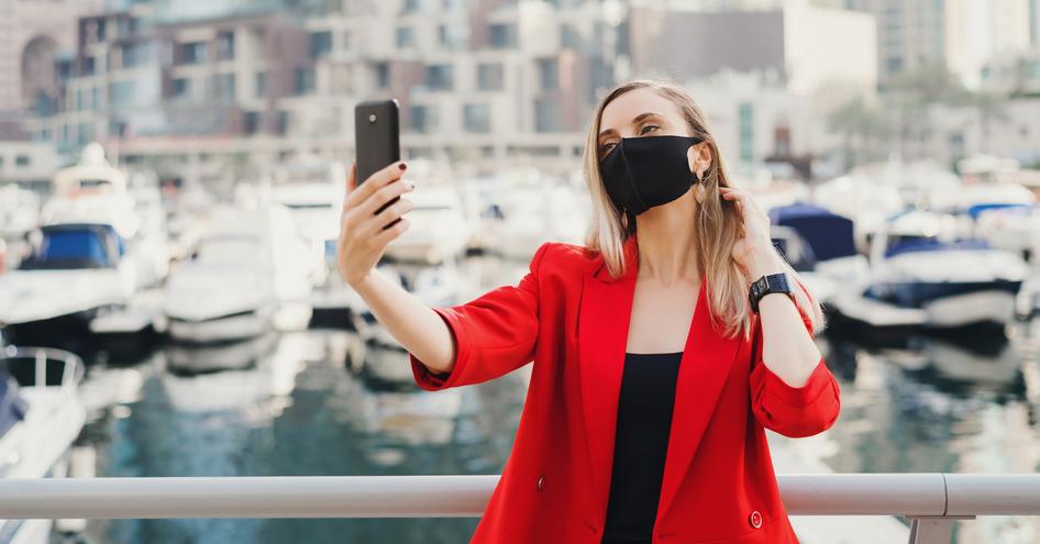 Woman in red suit posing for selfie in face mask, with multiple motor yachts moored in background.