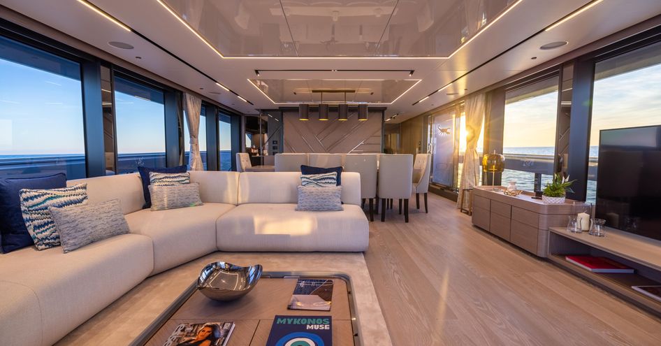 Main salon onboard Sunseeker 100 Yacht, white L shape sofa to port side, facing flatscreen TV. Surrounded by multiple large windows on both sides.