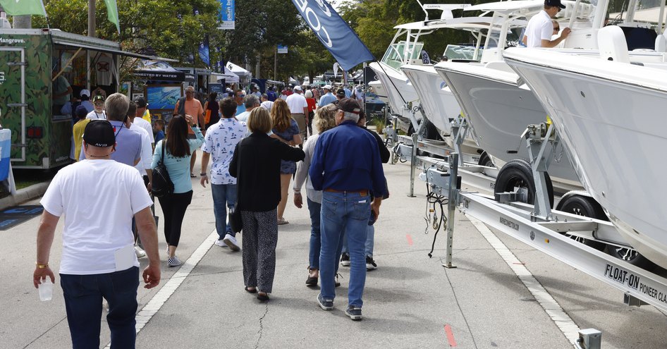 Steady stream of visitors walking along dock at Palm Beach International Boat Show, bows of superyachts just visible to starboard side.