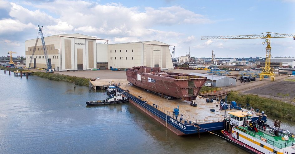 Oceanco shipyard with hull of boat leaving on floating barge. Crane and warehouses in the background