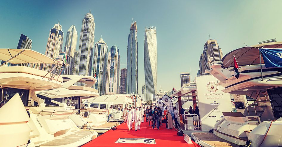 Overview at ground level of Dubai boat show, looking up towards skyscrapers, with yachts berthed either side of red carpeted slip.