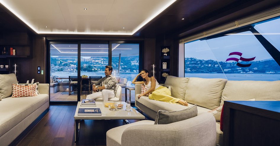 Main salon onboard Sirena Yachts 88, facing white sofas and coffee table with man and lady relaxing and watching TV