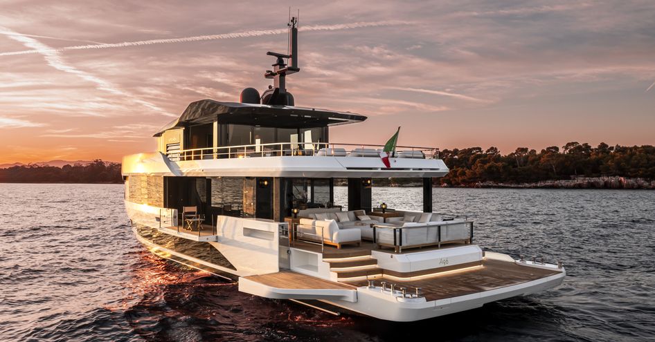 Arcadia A96 motor yacht shown at sunset, on anchor in still water with land in background; all side wings are open to extend the aft deck