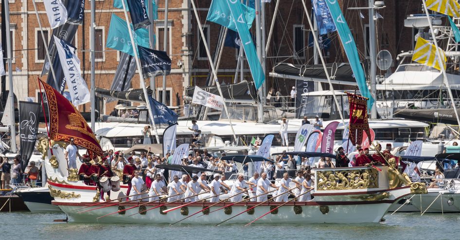 A regatta in action at the Venice Boat Show, with a wooden boat being rowed in the foreground and flags behind