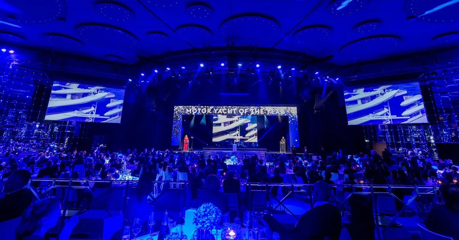 Overview of the World Superyacht Awards ceremony in 2021. Presenter on stage speaking to room full of tables and nominees. 